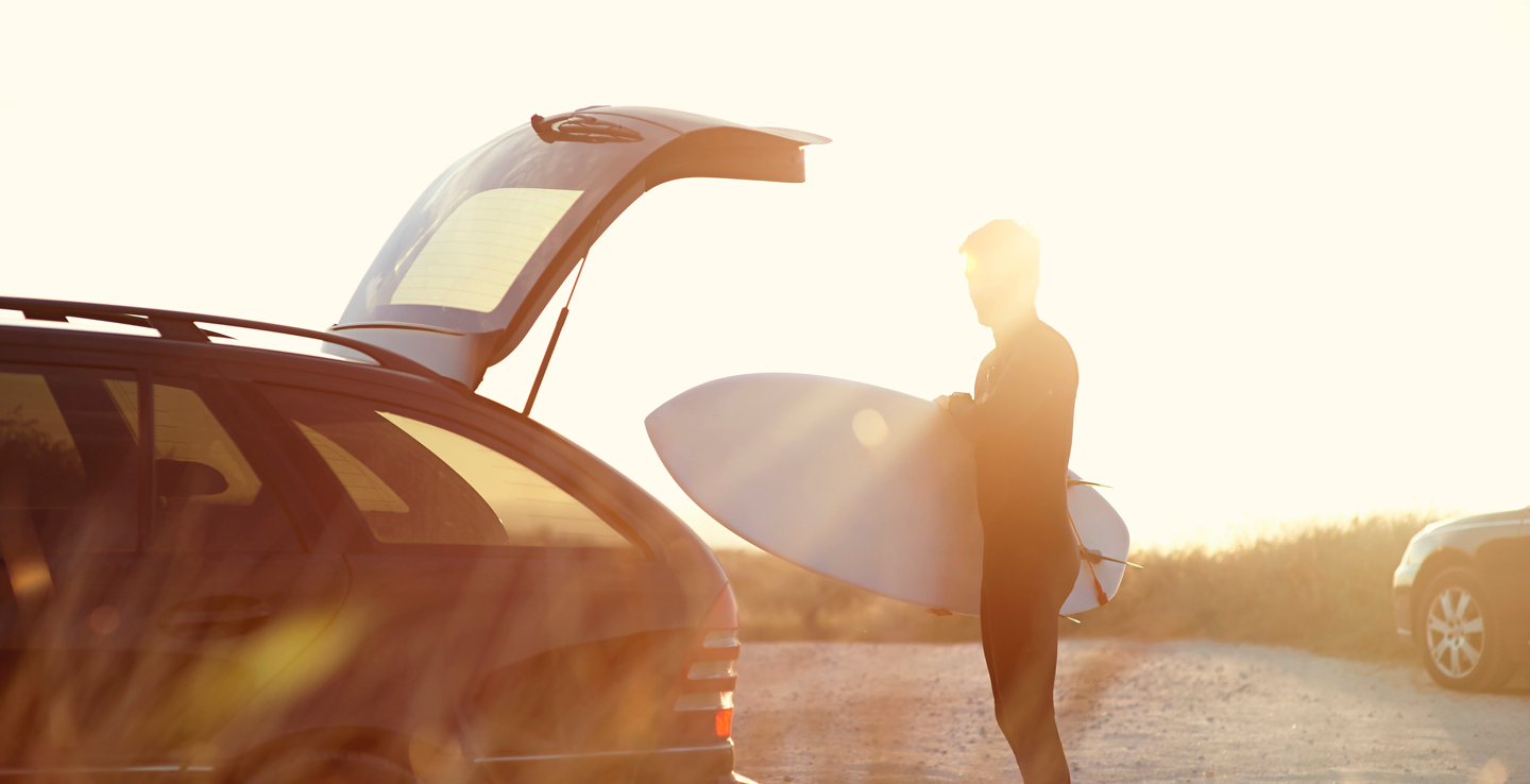 A man putting his surfboard in the boot of his station wagon around sunset.